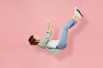 Full body side view young happy fun woman 20s she wear green shirt white t-shirt fly up hover over air fall down use mobile cell phone isolated on plain pastel light pink background studio portrait.