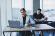 © Дмитрий Ткачук - A female manager is sitting at a desk using a laptop in a modern office against the background of a colleague. Working atmosphere in the office.