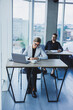 © Дмитрий Ткачук - A female manager is sitting at a desk using a laptop in a modern office against the background of a colleague. Working atmosphere in the office.