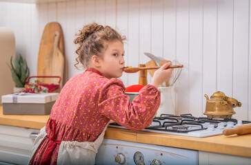 A little girl in the kitchen in an apron cooks something in a saucepan on a gas stove.