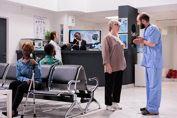 Wall Mural - Elderly woman receiving instructions from general practitioner in hospital waiting room. African american receptionist multitasking at clinic lobby. Consistent hospital emergency room.