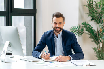 Photo of a positive successful handsome caucasian businessman, company ceo, manager, investor, wearing a suit, sits at a desk in the office, looking at the camera, smiles, taking notes in a notebook