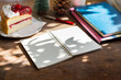 Sofe focus at blank note book with christmas cake, note book put on tablet computer, blurred small christmas tree and pine cone at background, Concept people relax in cafe after work on christmas