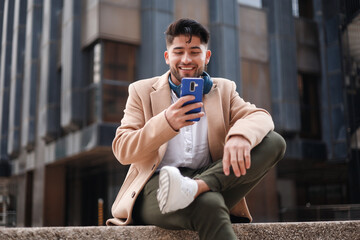 young stylish latin man using cellphone sitting outdoors