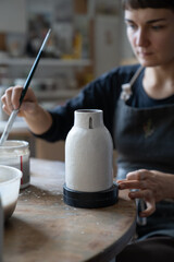 Poster - Carried away woman artist holding brush for applying traditional or creative patterns to ceramic liquid vessel. Soft focus female ceramist making tableware by herself in home pottery workshop