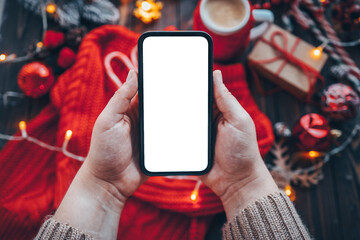 Women's hands in a warm knitted sweater with a smartphone holder with an isolated screen on Christmas background with gifts. Happy New Year and Merry Christmas