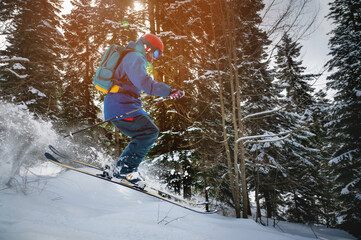 skier makes a ski jump with a plume of snow behind him, in the mountains against the backdrop of a dense forest. male athlete in bright clothes makes a downhill race