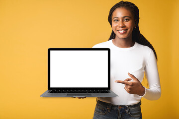 Satisfied multiracial woman advertising new computer app isolated on yellow background, mock-up. Positive smiling female freelancer holding and showing laptop with empty screen