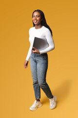 Young African American woman with afro hairstyle wearing smart casual wear and stylish eyeglasses standing isolated on yellow , carrying laptop computer, smiling, female office employee, student