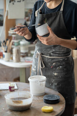 Poster - Creative craftswoman is engaged in manufacture of handmade ceramics standing near table in pottery studio. Cropped girl artist in apron holds brush and miniature vase makes dishes for souvenir shop