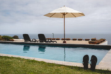 Flip-flops in the grass in front of a big pool in terrace with chairs and parasol and the ocean in a summer evening