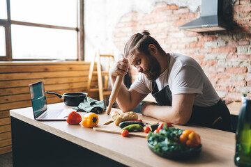 Bearded man watching video on laptop before cooking healthy dinner in kitchen