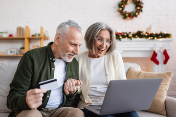 Wall Mural - happy mature man holding credit card near excited wife with laptop while having online shopping during christmas