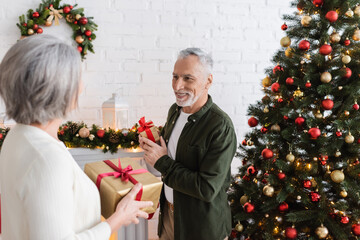 Wall Mural - smiling middle aged man holding present and looking at wife near christmas tree