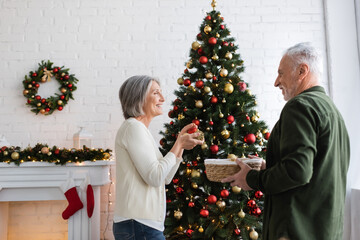 Wall Mural - smiling mature woman with grey hair decorating christmas tree and looking at husband holding wicker basket