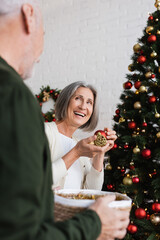 Wall Mural - joyful middle aged woman with grey hair decorating christmas tree near husband