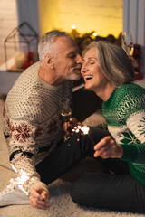 Wall Mural - positive middle aged man kissing wife and holding shiny sparklers on christmas eve