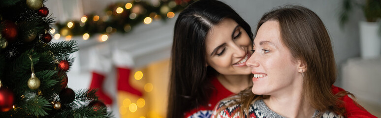 Wall Mural - Smiling woman kissing girlfriend in sweater near christmas tree at home, banner