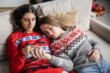 Sticker - Young same sex couple in christmas sweaters touching blurred hands on couch at home