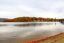 Lake View From The Beach With Fall Foliage Cliff And Houses On The Other Lakeside Under Cloudy Sky