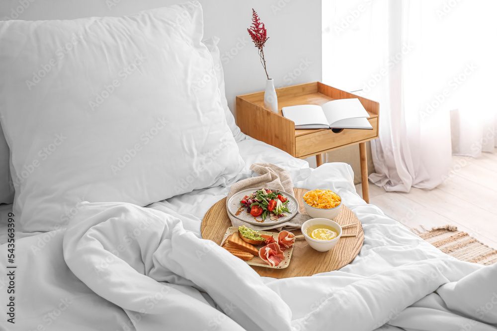 Board with tasty breakfast on white blanket in bedroom