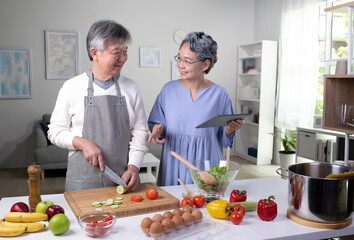 Wall Mural - Happy Asian senior couple is using a digital tablet, talking and smiling while cooking together in kitchen.