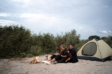 Romantic couple tourists laugh sitting at a campfire near tent, hugging each other under trees and night sky. Night camping. carefree couple relaxing remote travel