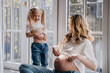 Mother and daughter at home in blue denim jeans and white t-shirts comparing their bellys. Pregnant mom shows her belly to little blonde girl with two tails. Maternity and childhood.