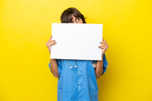 Young Nurse Doctor Woman Isolated On Yellow Background Holding An Empty Placard And Hiding Behind It