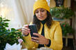 © Taras Grebinets - Close-up portrait of multi-ethnic pretty woman in bright yellow jacket and hat, using smartphone, checking news feed, scrolling web pages, checking social media content while chilling in a coffee shop