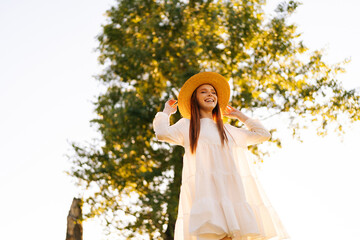 Wall Mural - low-angle view of laughing young woman with closed eyes in straw hat and white dress standing posing
