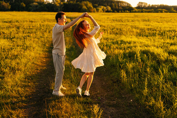 Wall Mural - cheerful young couple in love dancing together on green meadow in summer evening during golden sunse
