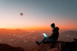 silhouette of digital nomad sitting on top of a hill working with his laptop over the city at dusk