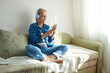 © evafesenuk - Mature woman sitting on sofa in headphones with smartphone