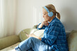 © evafesenuk - Senior woman in blue headphones sits on the couch at home, uses a mobile phone