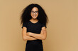 Slim satisfied woman with Afro haircut, wears black casual clothes, optical glasses, has confident expression, listens interlocutor, isolated on beige background. Cute teenage girl enjoys life.