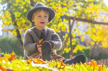 Little boy with hat sitting outside in grass during fall season having fun with leaves.