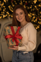 Wall Mural - A happy girl with red hair holds gift boxes near her face and smiles against the background of a Christmas tree and a garland. Close-up portrait of a girl for christmas. Merry Cristmas