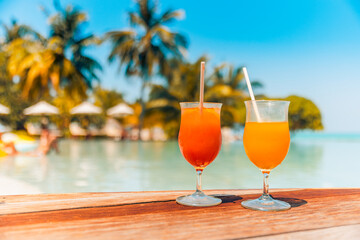 Cocktail glasses at summer poolside, resort beach side. Tropical island closeup outdoor bar cafe or restaurant with wooden table two cocktails, blurred umbrellas beds and calm swimming pool recreation