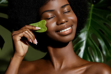 Wall Mural - gua sha jade skin treatment. african american model making face massage with a stone tool against tr