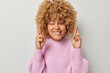 Photo of curly haired hopeful woman bites lips keeps fingers crossed believes in good luck makes wish winks eye wears casual pink knitted jumper isolated over grey background. Body language concept