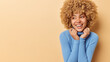 Indoor shot of sincere curly haired woman looks happy aside keeps hands on collar of turtleneck feels glad expresses positive emotions isolated over beige background copy space for your promotion