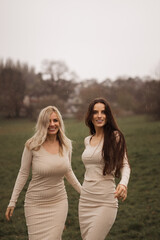 Two beautiful women sisters friends hugging, walking on green outdoors background. Life style, travel concept. Models under rain looking aside.