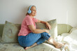 © evafesenuk - Smiling senior woman in headphones listening to music or audiobook while sitting on sofa at home