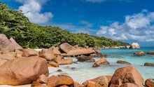 Picturesque Granite Boulders Are Scattered Along The Ocean Shore And In Turquoise Water. A Hill Covered With Green Tropical Vegetation, Against A Background Of Blue Sky And Clouds. Seychelles. Praslin