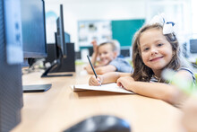 Smiling Girl With Book And Pencil Sitting At Desk In Classroom
