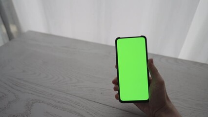 Wall Mural - Man showing and using smarphone with green screen while sitting behind white oak table