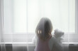 Little girl holding white teddy bear standing alone at window behind transparent day curtains and looking out from home. Rear view. Waiting concept. Close up.