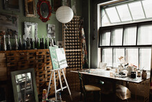 Art Workshop With Large Window And Vintage Furniture Near Bottles With Candles And Mirrors On Wall.