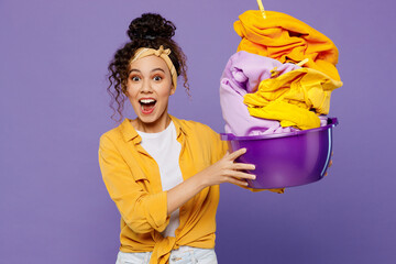 Young shocked happy surprised housekeeper woman wear yellow shirt tidy up hold show basin with clean clothes after laundry isolated on plain pastel light purple background studio. Housework concept.